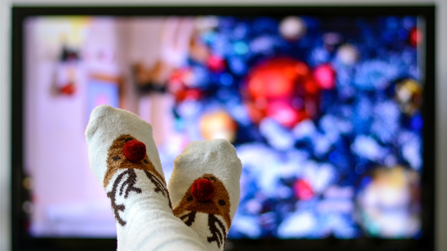 Person wearing reindeer socks lounges while tuning into a holiday movie.
